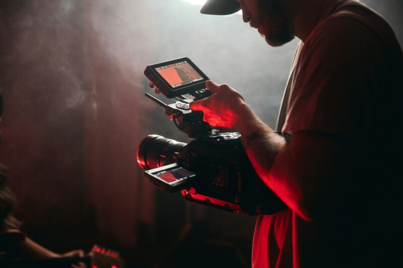 A close-up view of a person wearing a plaid shirt and red beanie holding and operating a professional black cinema camera and external monitor on a film set with hazy red and blue lighting.