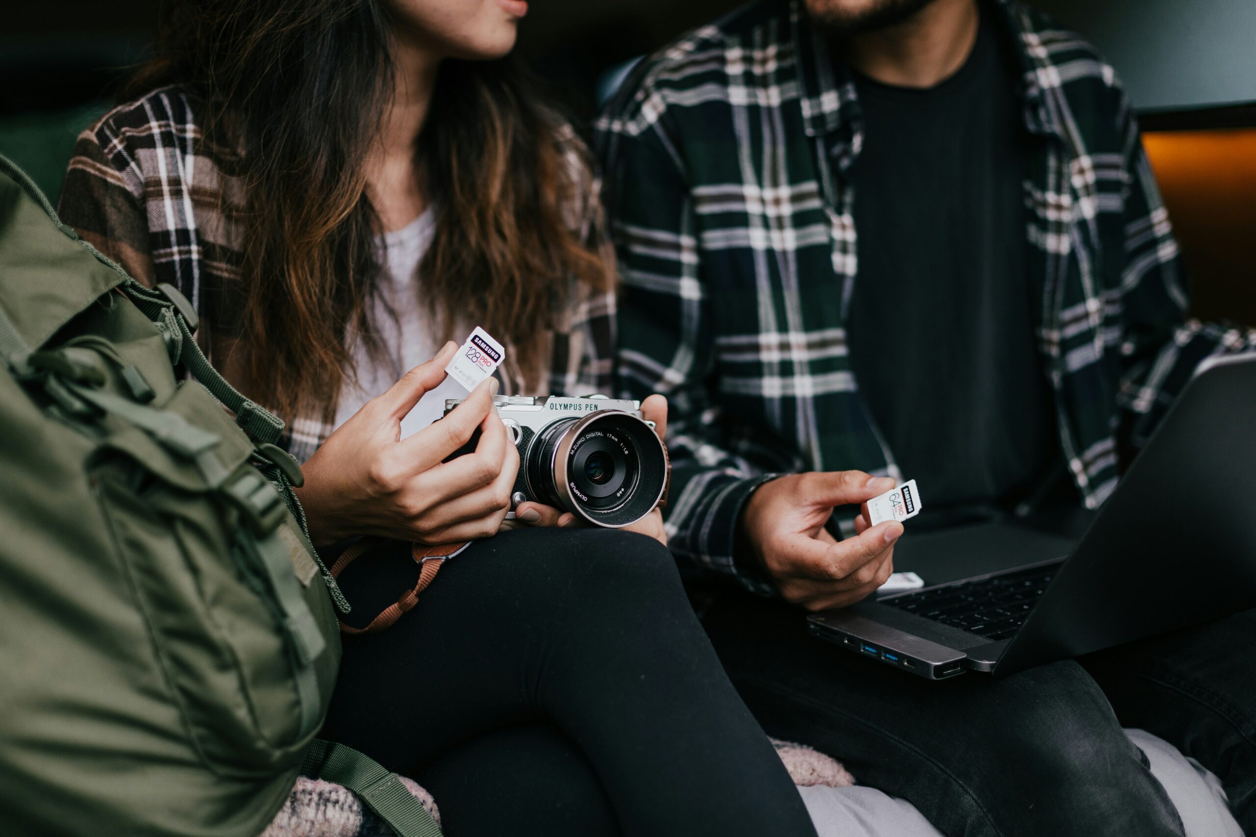 A pair of hands holding a camera and memory cards over a green backpack and laptop, representing creative digital content and social media post design by a marketing agency.