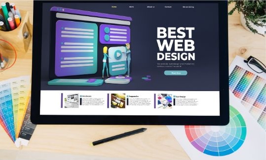 Top-down view of a digital workstation. A large tablet sits in the center, showing a website interface with a dark theme, 3D character illustrations, and bold white typography that reads "BEST WEB DESIGN." The desk is cluttered with creative professional tools, including color palette fans, printed color wheels, a black stylus, and a container of markers, suggesting a professional web development and digital marketing environment.