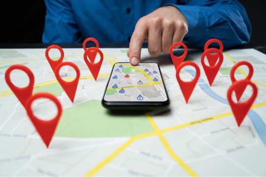 A conceptual image representing Local SEO. A person in a blue shirt is shown from the chest down, pointing at a map displayed on a smartphone. The phone is resting on a large, printed city map. Several oversized, red 3D location pins are digitally superimposed across the physical map, highlighting various points of interest and suggesting business visibility in a specific geographic area.