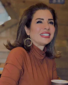 A professional lifestyle portrait of a smiling female physician with styled brown hair, wearing a warm brown turtleneck sweater and large decorative gold earrings in a soft-focused, cozy indoor setting, highlighting high-quality personal branding and authentic content creation for medical practitioners by MetaBaz.